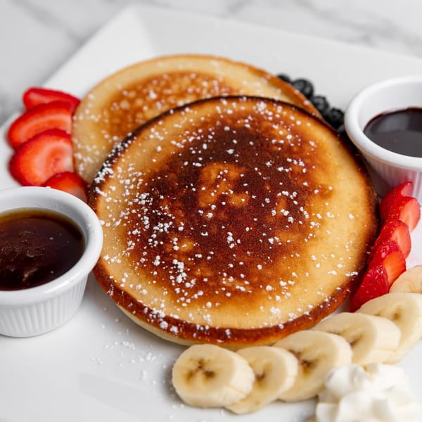 Golden pancakes with powdered sugar, sliced strawberries, bananas, and syrup on a white plate.