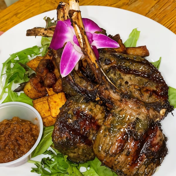 Grilled lamb chops with purple orchids, sweet potatoes, and a side of sauce on a bed of greens.